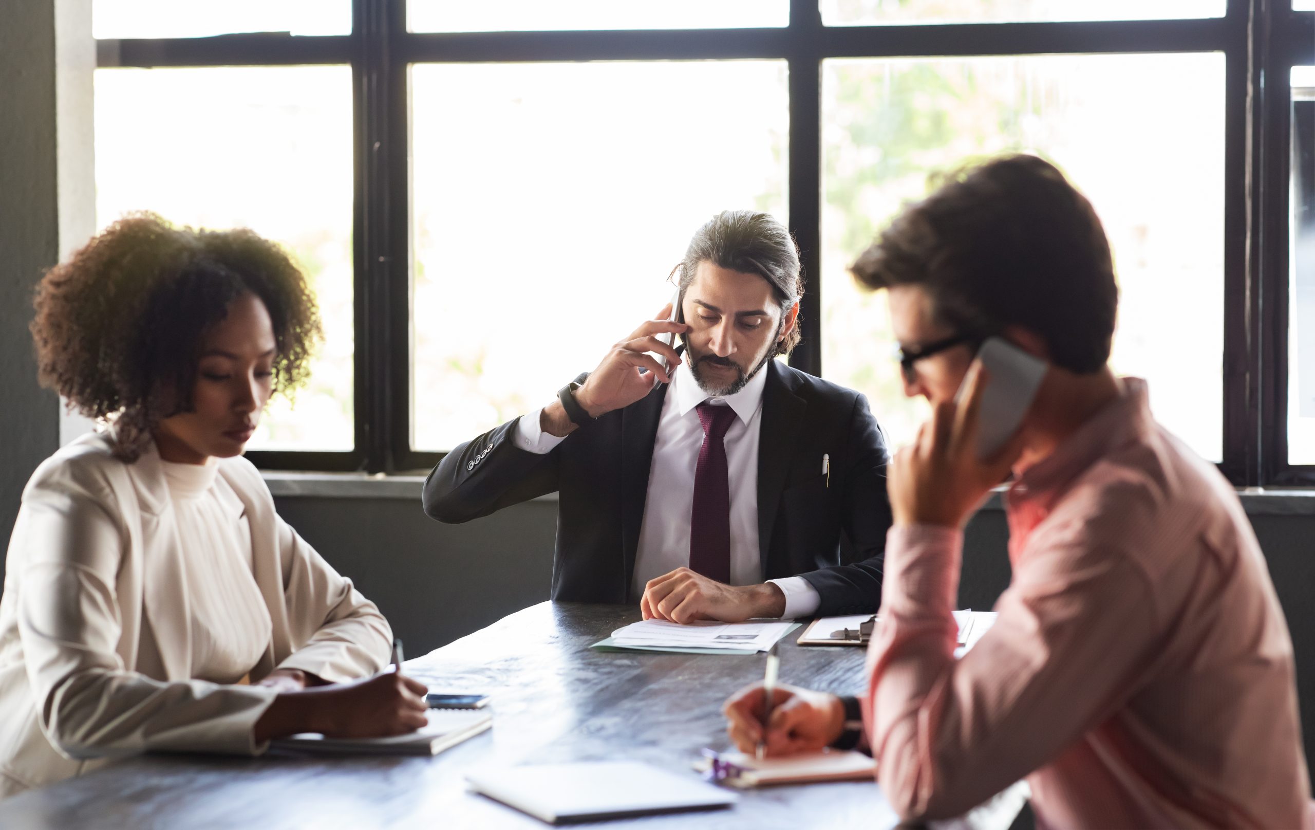Teamwork, collaboration, and cooperation. Multicultural team sitting at conference room against window at modern office, have morning briefing, work meeting, talking on smartphone, taking notes